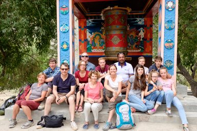 Eine Reisegruppe sitzt lächelnd vor einem bunten tibetischen Tempel mit einem großen Gebetsrad im Hintergrund.