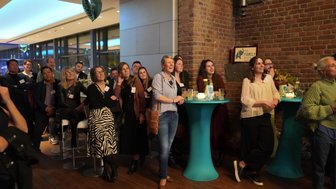 Eine Gruppe von Menschen steht in einem modernen Raum, aufmerksam und lächelnd, während sie einer Präsentation lauschen.