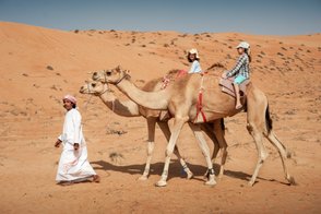 Kinder reiten gemeinsam mit einem Einheimischen auf Kamelen – Oman Reise mit Kindern