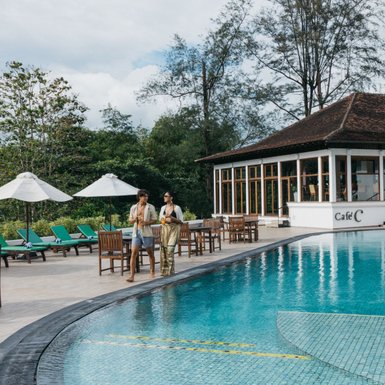 Ein Paar schlendert am Rand eines glitzernden Pools, umgeben von grünen Liegen und Sonnenschirmen, unter einem blauen Himmel.