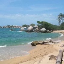 Ein ruhiger Strand mit sanften Wellen, umgeben von großen Felsen und üppigen Palmen im Hintergrund.