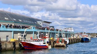 Am Hafen sind mehrere Fischerboote festgemacht, während eine Menschenmenge auf der Promenade steht und zuschaut.