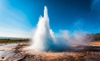 Ein beeindruckender Geysir sprudelt in die Höhe, umgeben von dampfenden Quellen und einer klaren blauen Himmel.