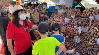 Eine Frau mit Sonnenhut und Maske steht an einem Marktstand, während zwei Kinder interessiert die Kartoffeln betrachten.