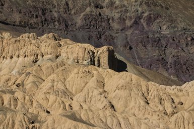 Die sanften, sandfarbenen Hügel erstrecken sich unter einem klaren Himmel, während die Felsen im Hintergrund in dunklen Tönen leuchten.