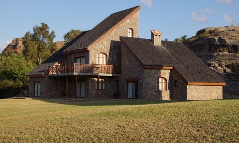 Ein modernes Steinhaus mit einem schrägen Dach steht in einer grünen Wiese, umgeben von sanften Hügeln und Bäumen.