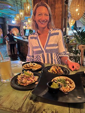 Eine lächelnde Frau sitzt an einem Tisch in einem Restaurant, umgeben von köstlichen mexikanischen Gerichten und Getränken.