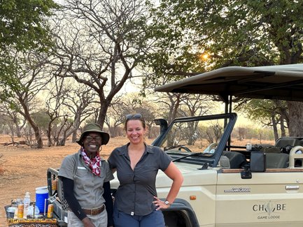 Zwei Personen stehen lächelnd vor einem Geländewagen in der afrikanischen Savanne, umgeben von kahlen Bäumen und sanftem Licht.