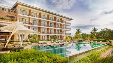 Ein modernes Resort mit mehreren Etagen, das von üppigem Grün und einem klaren Pool umgeben ist, unter einem strahlend blauen Himmel.