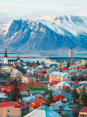 Blick auf Reykjavik mit bunten Häusern, umgeben von schneebedeckten Bergen und einem ruhigen Wasser im Hintergrund.