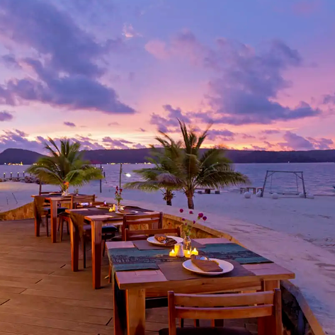 Ein romantisches Restaurant am Strand mit Holztischen, umgeben von Palmen und einem atemberaubenden Sonnenuntergang im Hintergrund.