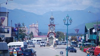 Die belebte Straße in Hokitika zeigt eine Uhr mit Glockenturm, umgeben von Geschäften und Bergen im Hintergrund.