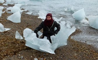 Eine Person sitzt auf einem großen Eisblock am Ufer, umgeben von kleineren Eisstücken und einem ruhigen Wasserhorizont.